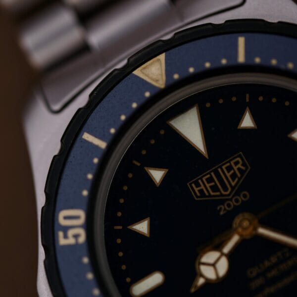 Close-up of a 1986 Tag Heuer 2000 Series Blue Bezel Ref. 972.606 1st Gen, showing part of the dark dial, luminous hands and markers, plus a section of the metal case and bezel with the number 50 visible.