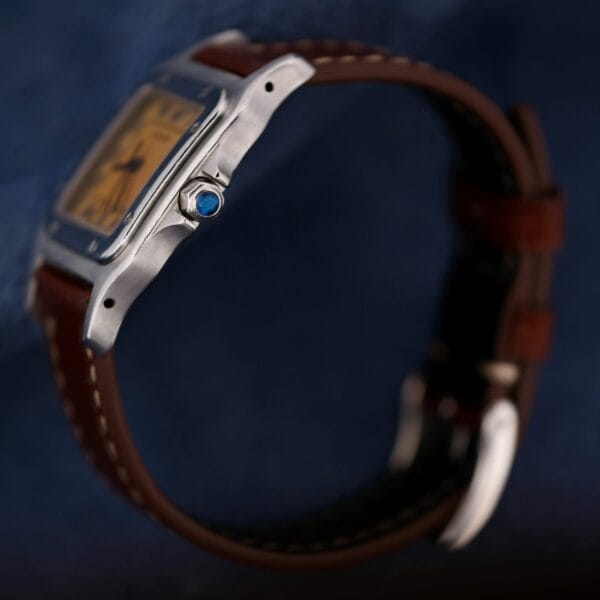 A close-up side view of the 1990s Cartier Santos Galbée ref. 1564 wristwatch with a rectangular silver case, brown leather strap, and a blue gemstone on the crown, set against a dark blue background.