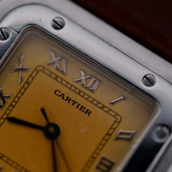 Close-up of a 1990s Cartier Santos Galbée ref. 1564 watch face featuring Roman numerals, yellow dial, and metal frame. The hour and minute hands are visible, with the Cartier name displayed at the center.