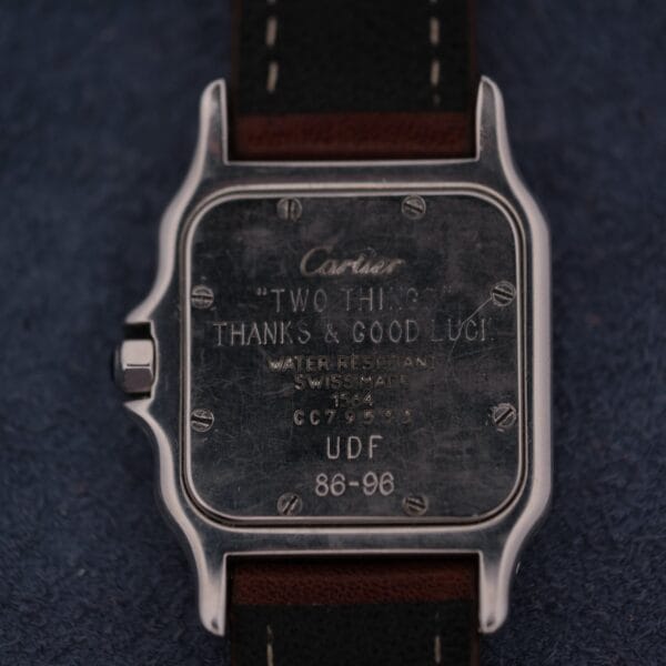 A close-up of the back of a 1990s Cartier Santos Galbée ref. 1564 with a brown leather strap. The engraving reads: “‘TWO THINGS. THANKS & GOOD LUCK!’ WATER RESISTANT SWISS MADE 1504 CC7964 UDF 86-96.”.