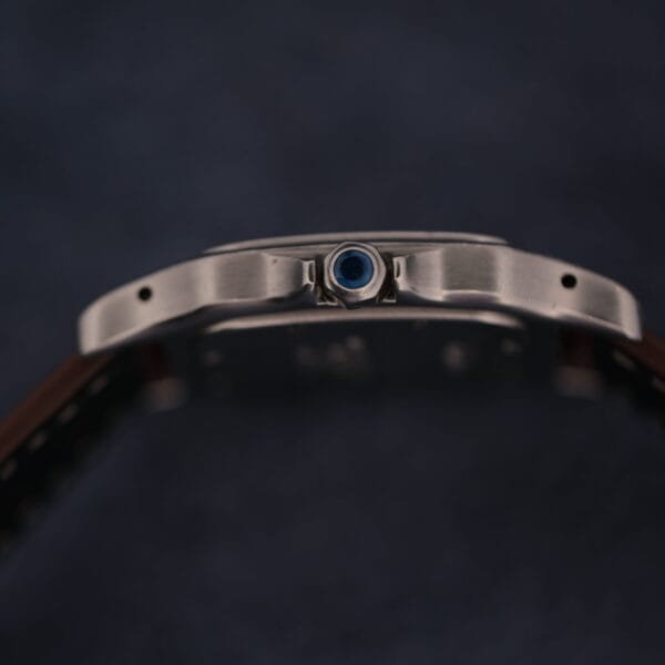 Side view of a 1990s Cartier Santos Galbée ref. 1564 case with blue cabochon crown and leather strap, set against a dark, blurred background. The metal watch case highlights the classic Cartier design.