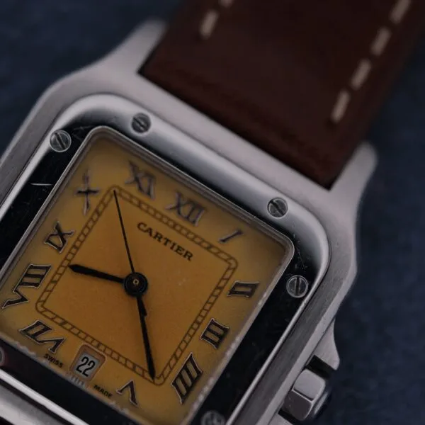 Close-up of a 1990s Cartier Santos Galbée ref. 1564 wristwatch featuring a square yellow-gold face, Roman numeral markers, black hands, date window at 6 o’clock, and brown leather strap with white stitching.
