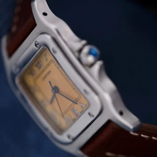 A close-up of a 1990s Cartier Santos Galbée ref. 1564 wristwatch with a brown leather strap, silver case, and yellowed Roman numeral dial. The crown is set with a blue gemstone against a dark, blurred background.