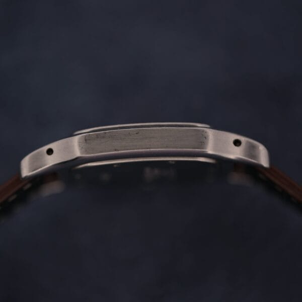 A close-up side view of a 1990s Cartier Santos Galbée ref. 1564 wristwatch case with a brushed metal finish, two visible screw holes, and a brown strap against a dark blurred background.