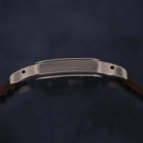 A close-up side view of a 1990s Cartier Santos Galbée ref. 1564 wristwatch case with a brushed metal finish, two visible screw holes, and a brown strap against a dark blurred background.