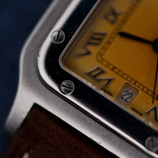 Close-up of a 1990s Cartier Santos Galbée ref. 1564, featuring a square metal case, yellow dial with Roman numerals, date window, visible bezel screws, and brown leather strap in focus.