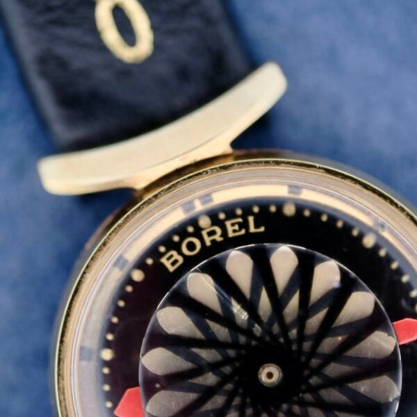 Close-up of the Ernest Borel Kaleidoscope Cocktail Black Dial watch, featuring a captivating kaleidoscope design with concentric circles and geometric patterns, a gold-colored case paired with a black leather strap, set against a textured blue background.