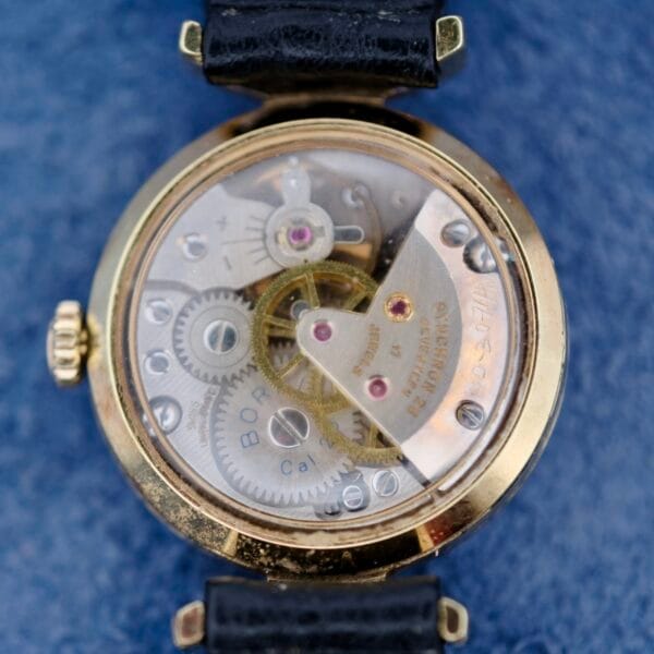 Close-up of an Ernest Borel Kaleidoscope Cocktail Black Dial watch with an open back case revealing visible gears. The watch features a gold-colored rim and black strap, resting on a blue textured surface.