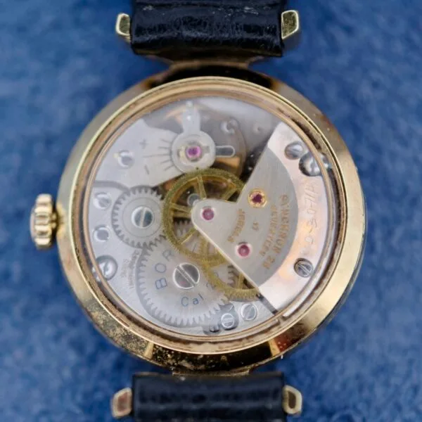 Close-up of an Ernest Borel Kaleidoscope Cocktail Black Dial watch with an open back case revealing visible gears. The watch features a gold-colored rim and black strap, resting on a blue textured surface.
