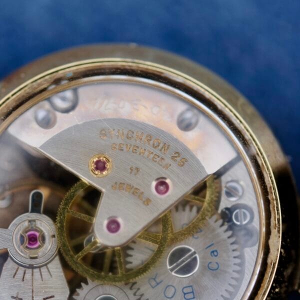 Close-up of a mechanical watch movement akin to the Ernest Borel Kaleidoscope Cocktail Black Dial, showcasing gears with SYNCHRON 26 and 17 JEWELS inscriptions. Gold-toned gears with red accents contrast against a blurred blue background.
