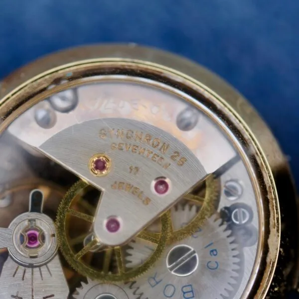 Close-up of a mechanical watch movement akin to the Ernest Borel Kaleidoscope Cocktail Black Dial, showcasing gears with SYNCHRON 26 and 17 JEWELS inscriptions. Gold-toned gears with red accents contrast against a blurred blue background.