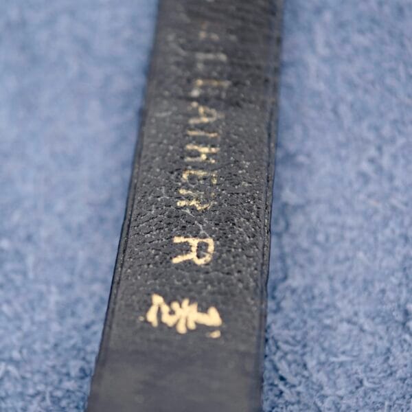 A close-up shows a black belt with white embroidery on a blue fabric background, reminiscent of an Ernest Borel Kaleidoscope Cocktail Black Dial. The belt displays a partially visible word or name with small symbols or characters, and the texture of both elements is clearly visible.