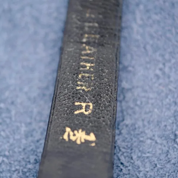 A close-up shows a black belt with white embroidery on a blue fabric background, reminiscent of an Ernest Borel Kaleidoscope Cocktail Black Dial. The belt displays a partially visible word or name with small symbols or characters, and the texture of both elements is clearly visible.