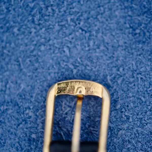 A close-up of an Ernest Borel belt buckle against a textured blue background highlights the gold buckles kaleidoscope-like reflective finish and simple rectangular design, exuding elegance and charm.