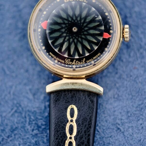 A vintage Ernest Borel Kaleidoscope Cocktail watch with a black dial and geometric pattern features two red hands. It has a gold case with a right-side crown and a black leather strap adorned with a gold design, all set against a blue fabric background.