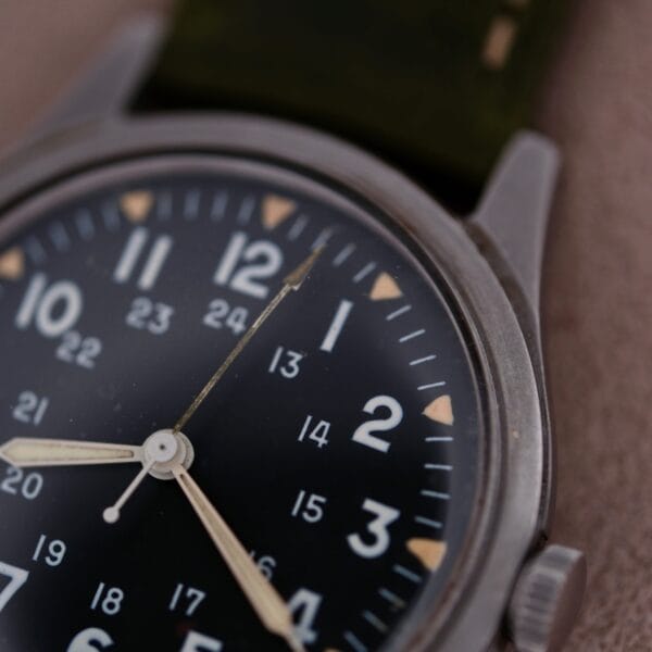 Close-up of the 1964 Benrus U.S. Military Issued DTU-2A/P wristwatch with a black dial, featuring 12-hour and 24-hour markings, housed in a sleek metal case with the Hack Second function. It has two white hands and a partially visible dark green strap against a soft-focus backdrop.