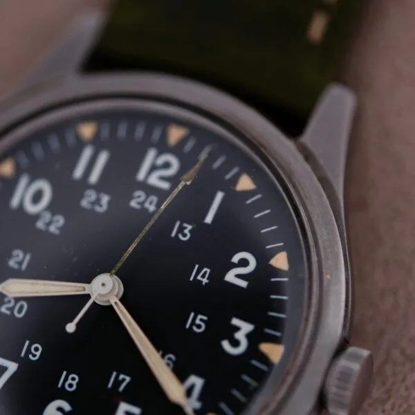 Close-up of the 1964 Benrus U.S. Military Issued DTU-2A/P wristwatch with a black dial, featuring 12-hour and 24-hour markings, housed in a sleek metal case with the Hack Second function. It has two white hands and a partially visible dark green strap against a soft-focus backdrop.