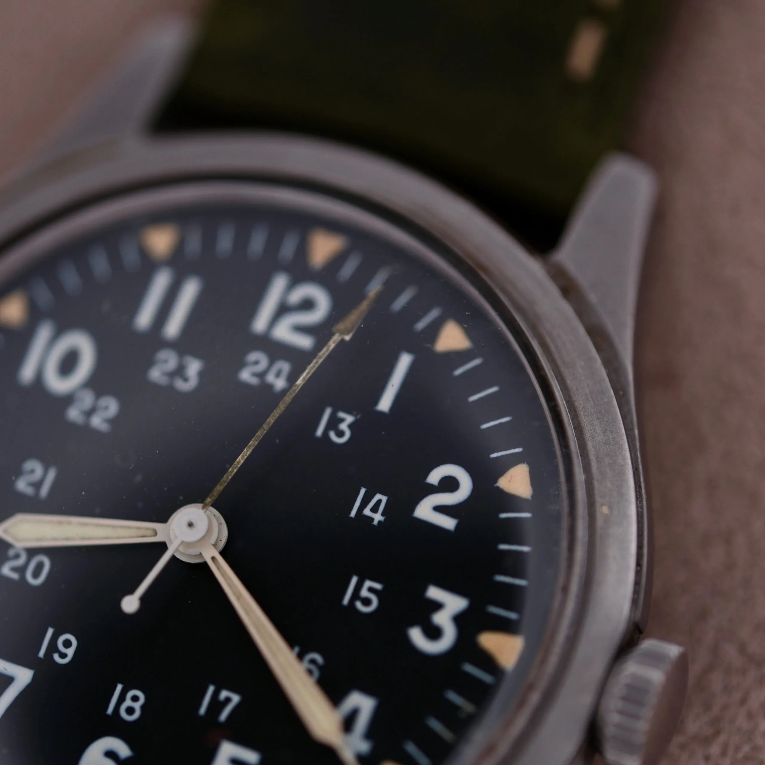 Close-up of the 1964 Benrus U.S. Military Issued DTU-2A/P wristwatch with a black dial, featuring 12-hour and 24-hour markings, housed in a sleek metal case with the Hack Second function. It has two white hands and a partially visible dark green strap against a soft-focus backdrop.