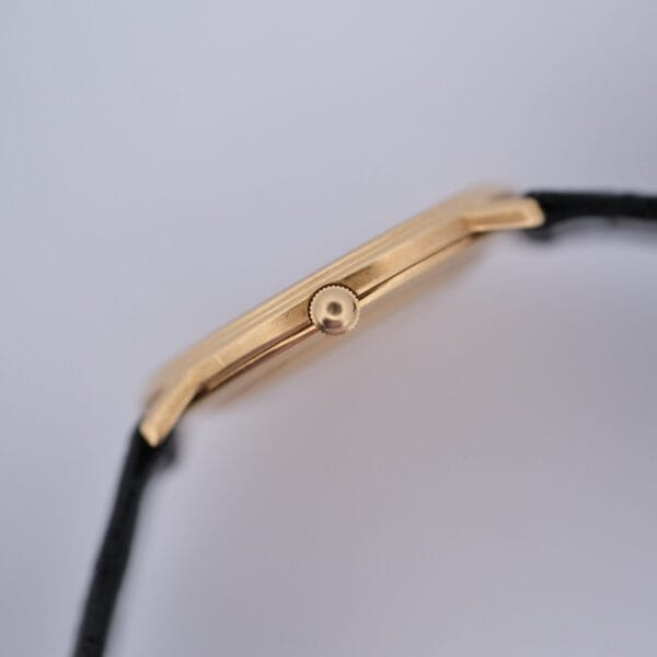 Side view of the Audemars Piguet Classic 18k Gold Roman Numerals watch case with a black strap and central round crown; watch face hidden, plain blurred background.