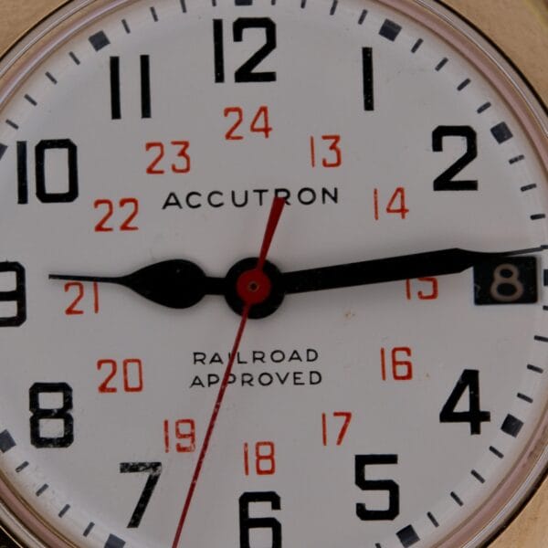 Close-up of a Bulova Accutron Railroad Approved Date clock face showing 8:14:32, with black hour/minute hands, a red second hand, black hour markers, red 24-hour numbers, and ACCUTRON and RAILROAD APPROVED text.