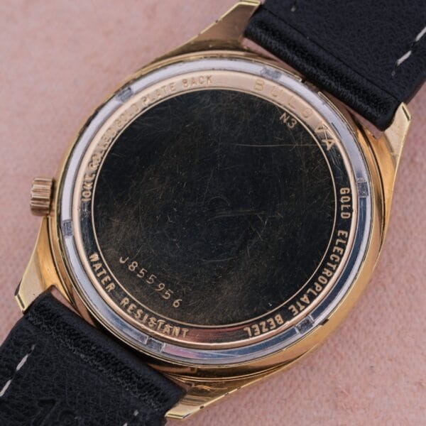 Close-up of the back of a Bulova Accutron Railroad Approved Date wristwatch with a gold-colored case, black leather strap, and engraved markings: N3, WATER RESISTANT, and serial number J855956.