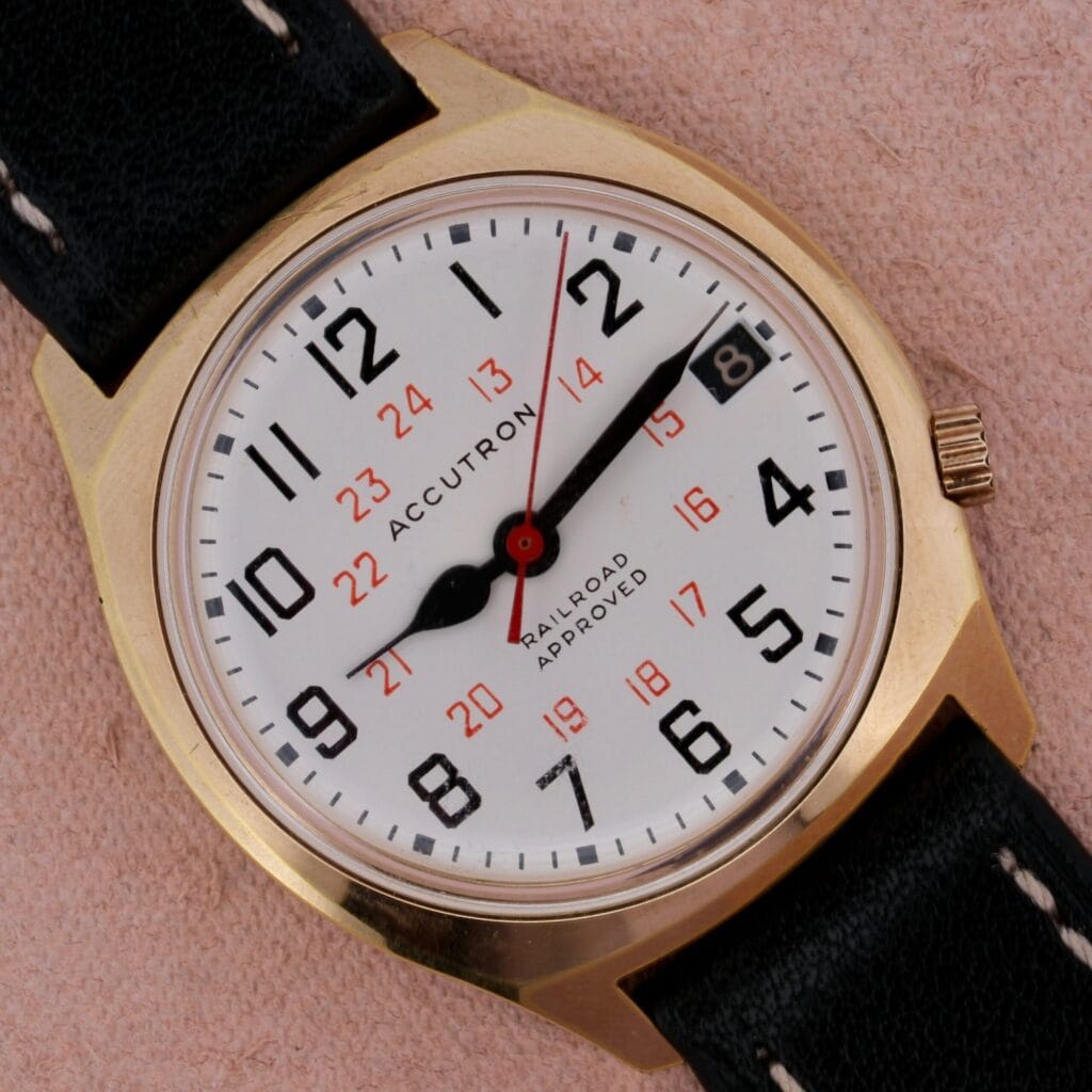 Gold Bulova Accutron Railroad Approved Date watch with black band, white dial, black numerals, red 24-hour markers, and date display.