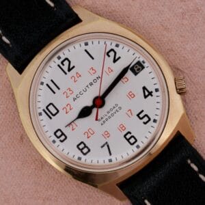 Gold Bulova Accutron Railroad Approved Date watch with black band, white dial, black numerals, red 24-hour markers, and date display.