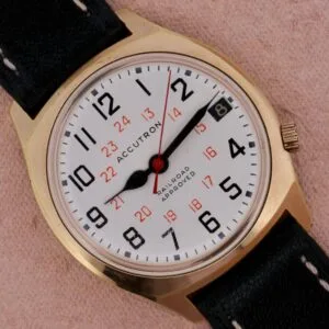 Gold Bulova Accutron Railroad Approved Date watch with black band, white dial, black numerals, red 24-hour markers, and date display.