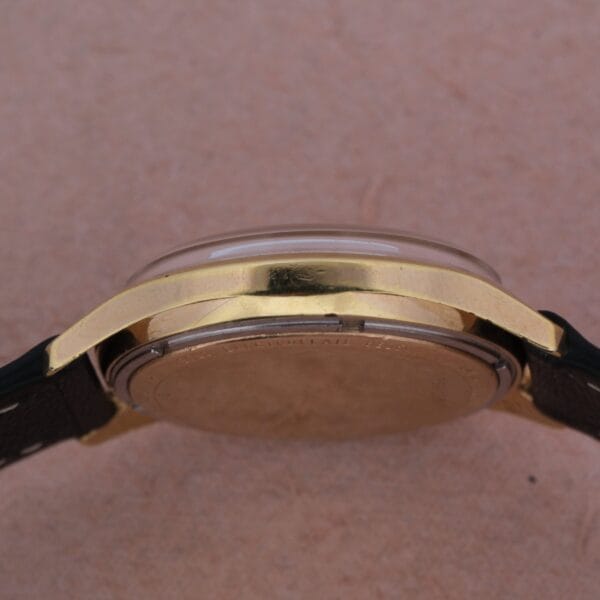 A close-up side view of the Bulova Accutron Railroad Approved Date wristwatch, featuring a gold-colored case, transparent crystal, and black leather strap against a plain brown background.