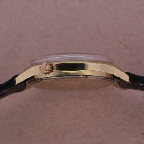 Side view of the Bulova Accutron Railroad Approved Date gold case with a black leather strap, displayed on a light brown textured surface. The engraved winding crown is visible on the left side of the watch.
