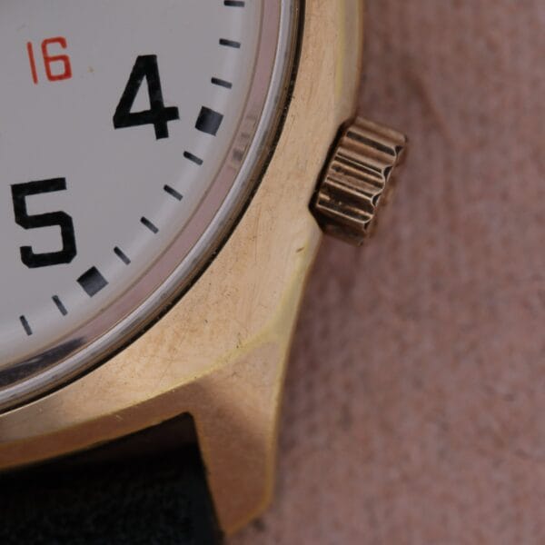 Close-up of the Bulova Accutron Railroad Approved Date wristwatch showing numbers 4 and 5 on a white face with black hour markers, an orange 16, visible metal casing and crown, and part of a dark strap.