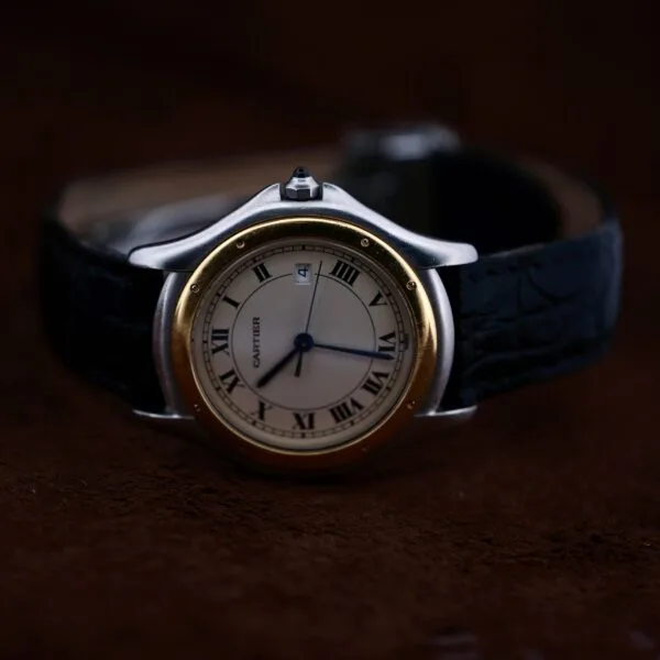 A close-up of the Cartier Cougar Gents Date Ref. 187904, featuring a round face, Roman numerals, blue hands, date window, gold bezel, and black leather strap against a dark brown background.