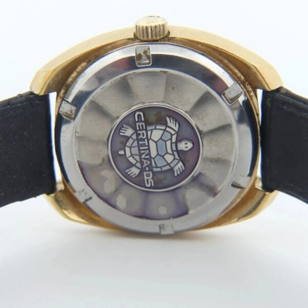 The image showcases the back of a Certina DS-2 Gold Turtle Case wristwatch. It features a turtle emblem centered on gold and silver-tone metal with an attached black strap on either side.