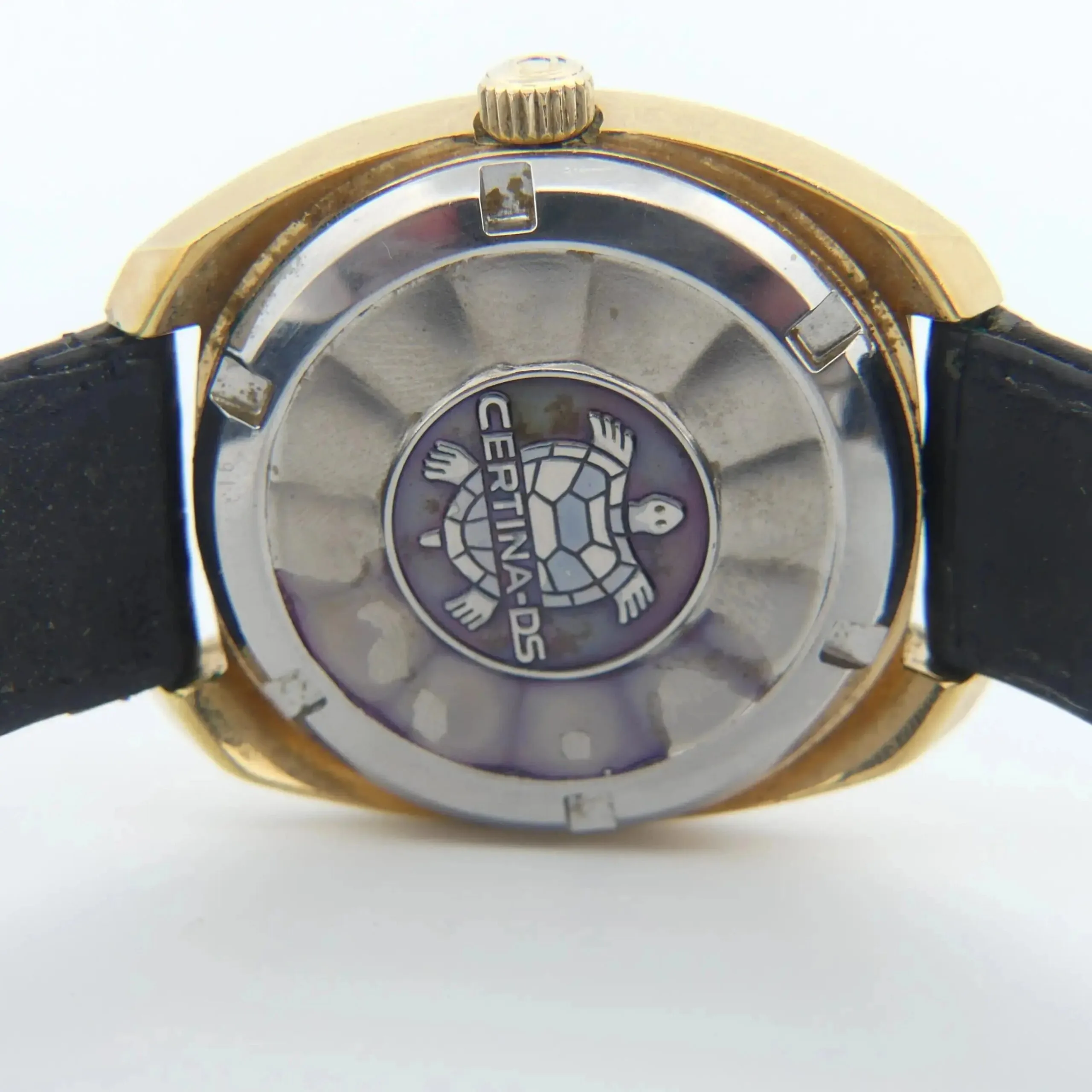 The image showcases the back of a Certina DS-2 Gold Turtle Case wristwatch. It features a turtle emblem centered on gold and silver-tone metal with an attached black strap on either side.