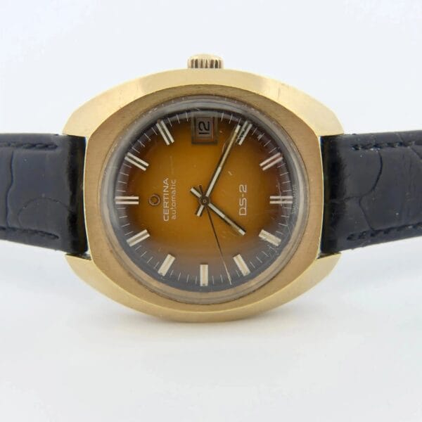 The Certina DS-2 Gold Turtle Case wristwatch features an orange-brown ombre dial, Cal. 25-651M movement, date at 3 o’clock, silver hour markers, and a black leather strap, displayed on a plain white background.