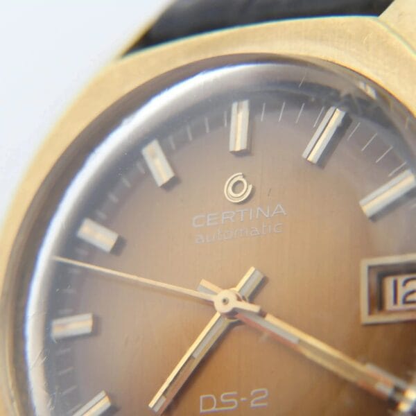Close-up of a Certina DS-2 Gold Turtle Case with an orange-brown ombre dial. The watch features silver hour markers, hands, and a date window on the right. The Certina logo sits below the 12 oclock position.