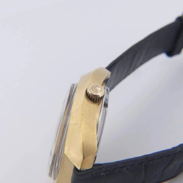 Side view of the Certina DS-2 Gold Turtle, featuring a gold case and round crown, with a black leather strap and an orange-brown ombre dial under a raised crystal. The background is light-colored and plain.