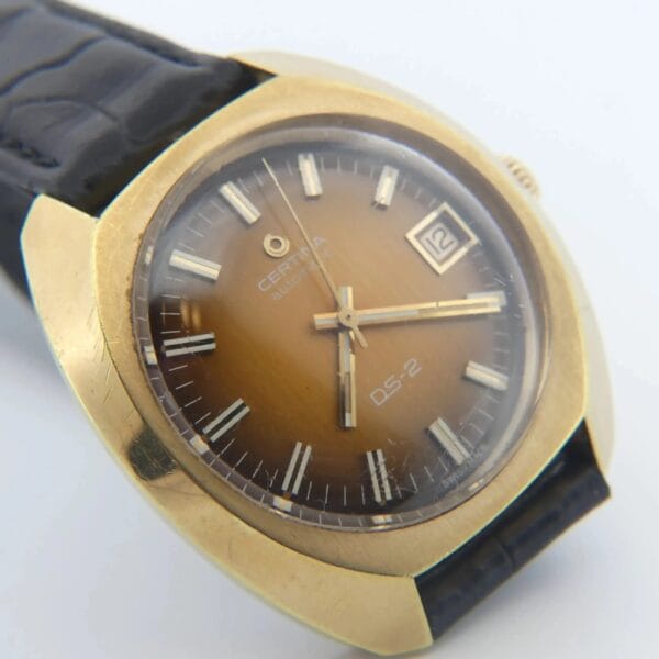 A Certina DS-2 Gold Turtle Case watch featuring an orange brown ombre dial, Cal. 25-651M movement, silver hour markers, date window at 3 o’clock, and a black textured leather strap.