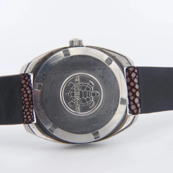 A close-up of the Certina DS-2 Turtle Case wristwatch showcases a black leather strap and silver case featuring an engraved turtle logo with CERTINA DS text. Near the case, the textured strap displays a dark brown section.