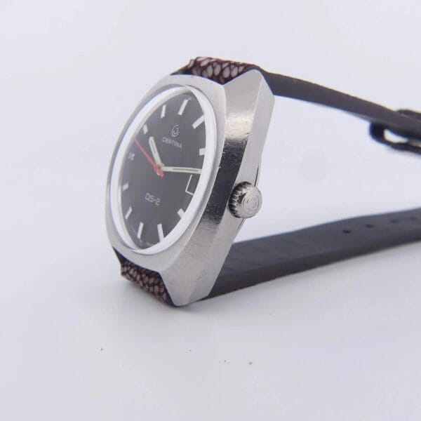 The Certina DS-2 Turtle Case displays a silver watch with a black dial, brown leather strap, white hour markers and hands, and a red second hand. Its minimalistic design includes a visible side crown.