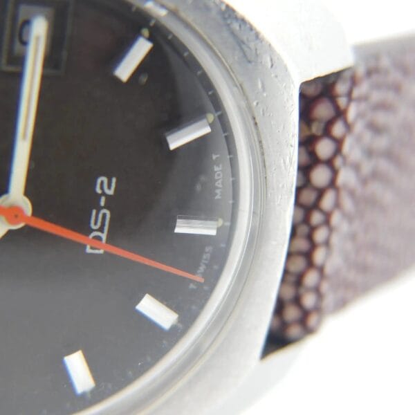 Close-up of a vintage Certina DS-2 wristwatch featuring a brown textured leather strap, black dial with white hour markers, and an orange second hand. Part of the metal case and T Swiss Made T is visible.