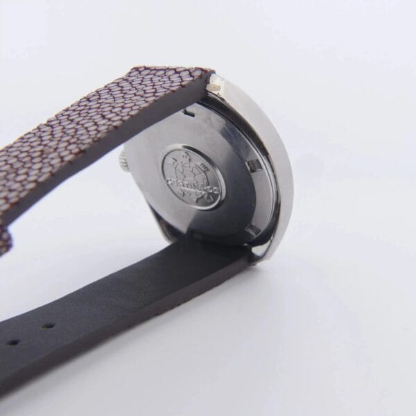 The image displays the back of the Certina DS-2 Turtle Case watch, featuring a silver case with a logo and text. The dark brown textured strap is partially lifted against a plain white background.