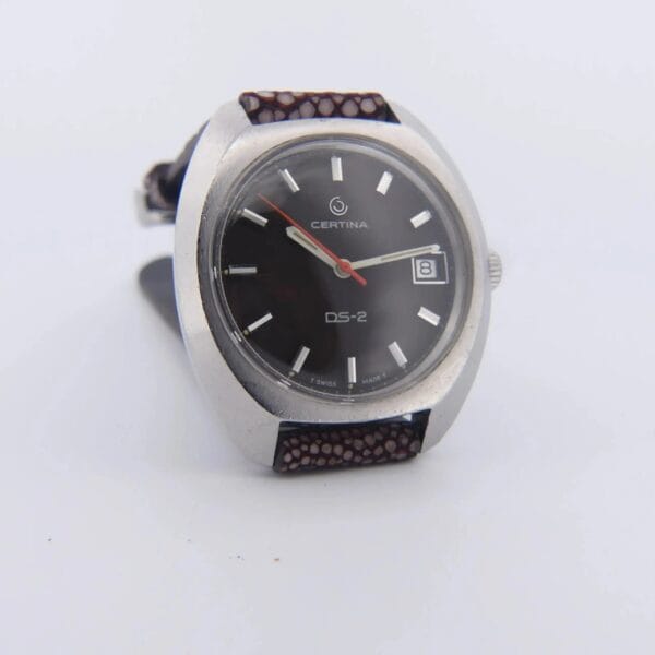 The Certina DS-2 Turtle Case Orange Second Black Dial Cal. 25-661 features a black dial with silver hour markers, a red-brown patterned strap, and a date display at 3 oclock within its circular silver case.