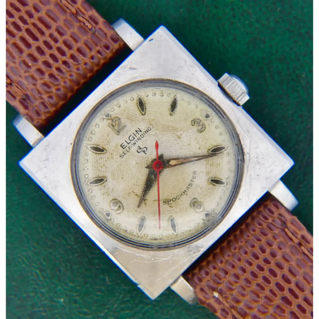 An Elgin Self-Winding Shock Master Square Case watch with a silver square case, cream dial, brown textured strap, and red second hand is displayed against a green background.