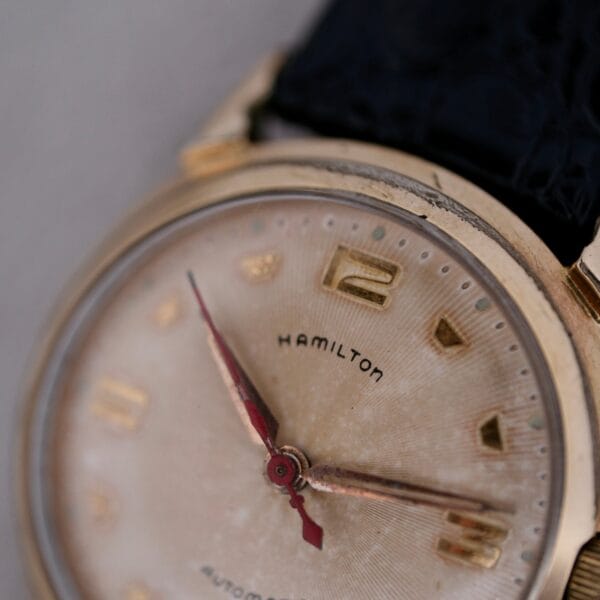 Close-up of a Hamilton Accumatic III Cal. 672 wristwatch with a gold-tone bezel, featuring gold hour markers at 3, 6, 9, and 12, three red hands, a worn look, and a textured black strap.