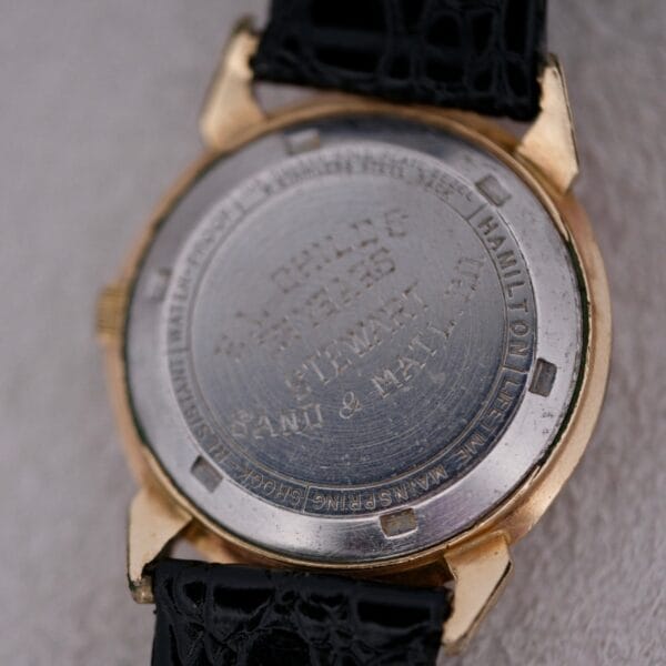 Close-up of the back of a Hamilton Accumatic III Cal. 672 with a gold case and black textured strap. The metallic back is engraved with text, though some parts are worn and hard to read. Watch is resting on a light surface.
