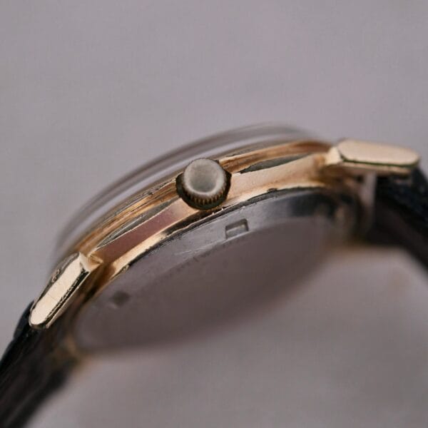 Close-up of the Hamilton Accumatic III Cal. 672, showcasing its gold-tone case and round crown. The edge of the watch face and a section of the black leather strap are visible, emphasizing the side profile of the metal casing.