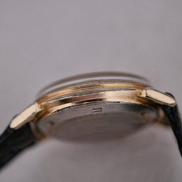 A close-up side view of the Hamilton Accumatic III Cal. 672 wristwatch features a gold-colored case and black leather strap. The watch face is partially visible, with a slightly raised glass.