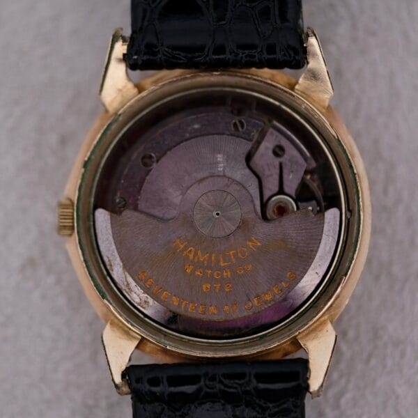A close-up of the back of a Hamilton Accumatic III Cal. 672 reveals visible inner mechanics, with Hamilton Watch Co. 672 Seventeen (17) Jewels on the mechanism. It features a black leather strap and a gold-colored case.