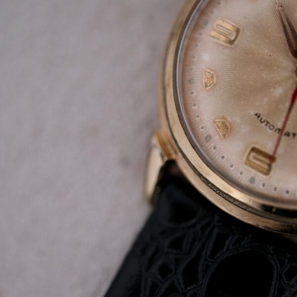A close-up of the Hamilton Accumatic III Cal. 672 wristwatch showcasing a gold case, black leather strap, and cream-colored face with gold hour markers and hands. The dial partially reveals the word Automatic.